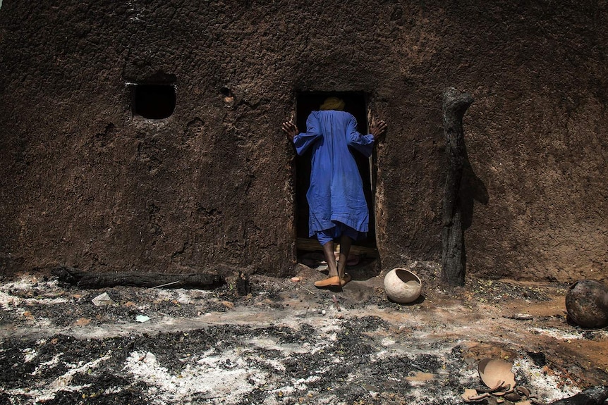 A person in a blue robe pictured from the back as they put their head in a charred doorway with ash on the ground.