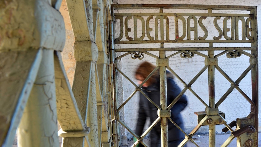 A blurred figure walks through metal gates that feature the motto "Jedem das Seine".