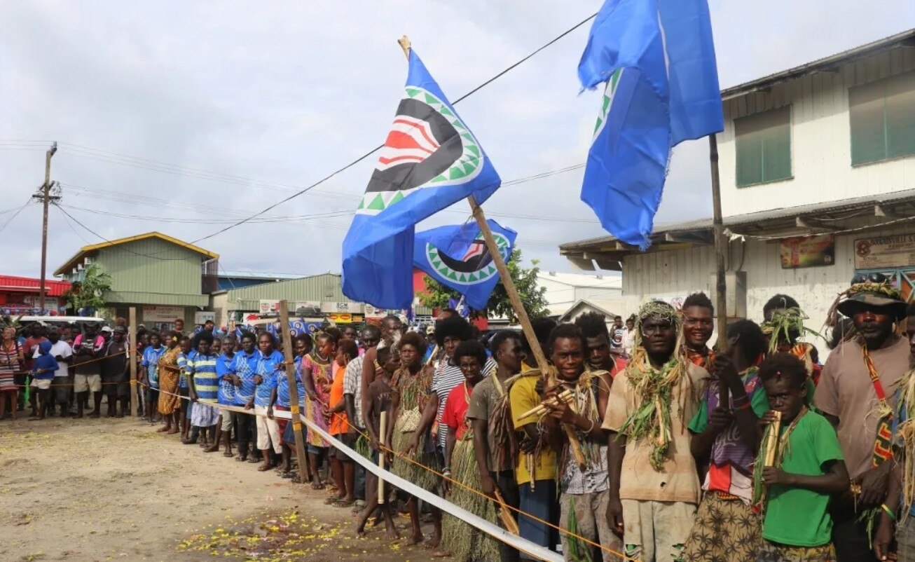 Large group of people line up outdoors to vote, some carry Bouganville flag. 