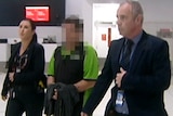 A man in working clothes and handcuffs is led through Perth Airport by two police officers.