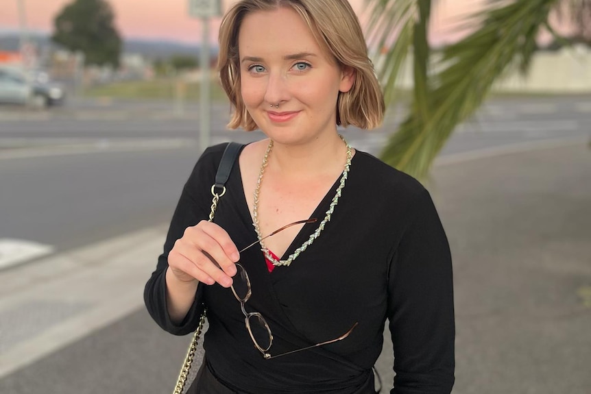 A young woman in black looks at the camera holding reading glasses, with a sunset in the background.