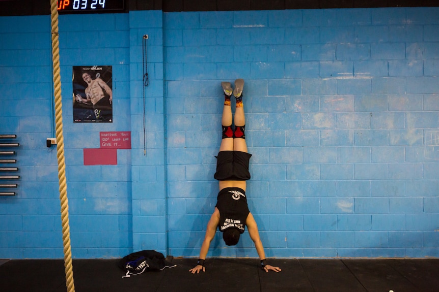 Father Justel Callos, in muscle shirt, shorts and knee supports, does a hand stand against a blue wall in his CrossFit gym.