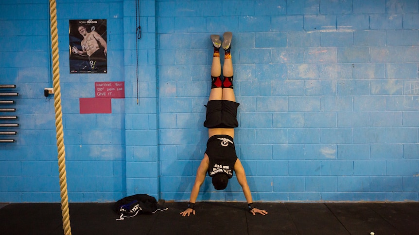 Father Justel Callos, in muscle shirt, shorts and knee supports, does a hand stand against a blue wall in his CrossFit gym.