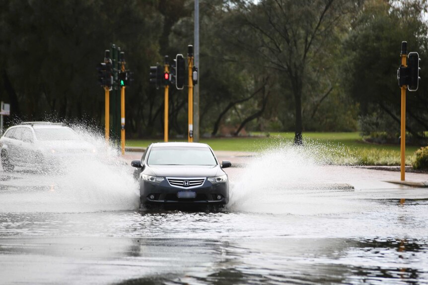 Large sprays of water shoot out from under a car as it drives on a flooded Perth street.