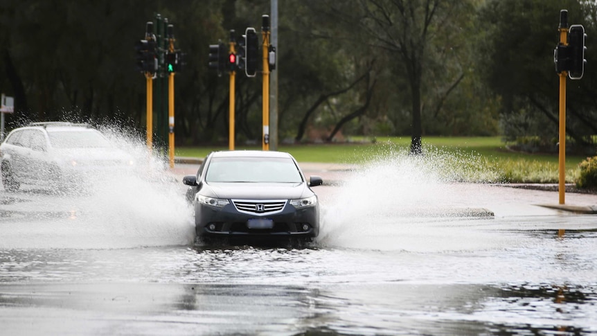 Large sprays of water shoot out from under a car as it drives on a flooded Perth street.