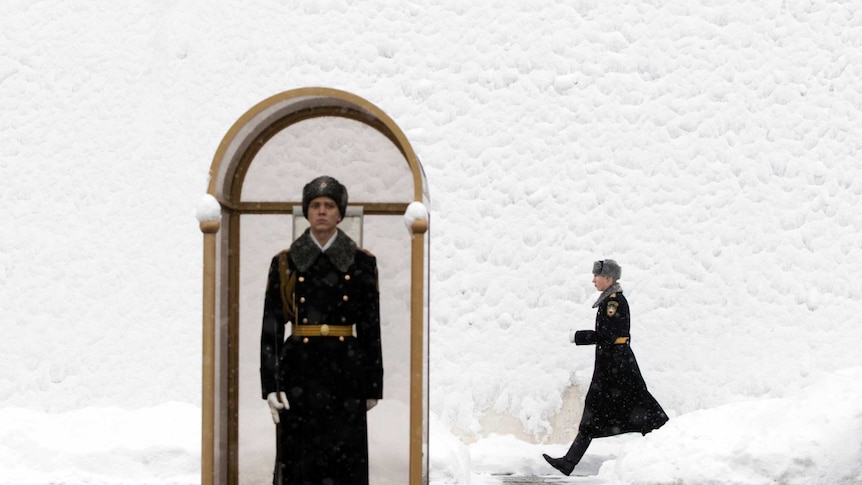 A Russian guard stands at his post outside snow covered Kremlin walls, a second guard marches behind in the snow.
