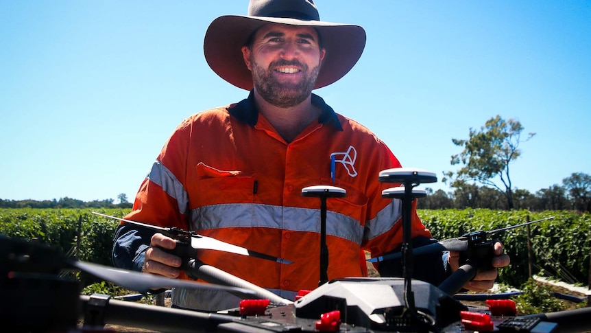 Drone pilot Luke Jurgens looks at the camera, he is wearing a big hat and high visibility shirt, standing with his drone.