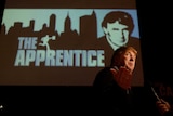 Donald Trump pointing and speaking at a podium in front of an Apprentice screen