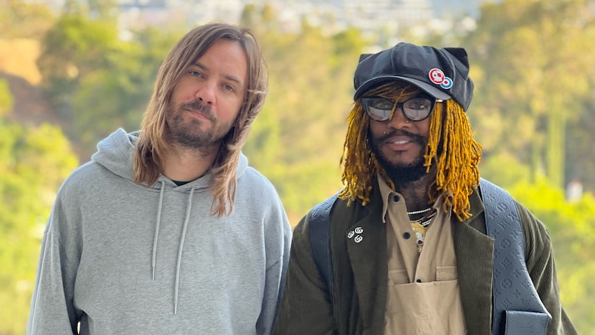 Kevin Parker in a grey hoodie with Thundercat in a beige shirt, blue overalls, dark green jacket and black leather vest.
