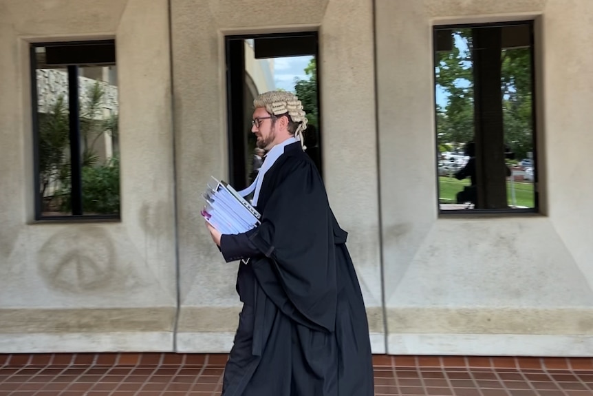 a man in a wig and black robes walks outside a cement building holding a stack of folders