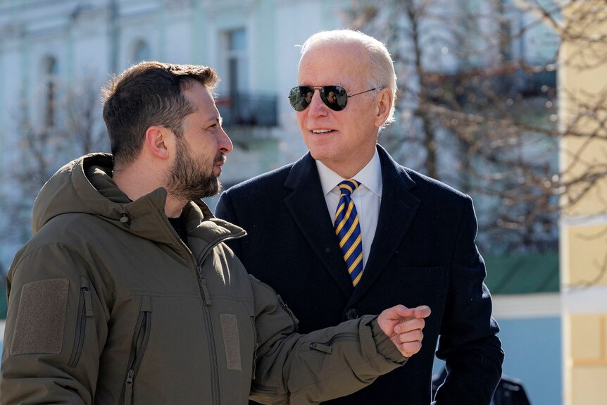 Zelensky gestures as he speaks, standing close to Biden, who looks overhead. Zelenskyy's face is reflected in Biden's sunglasses