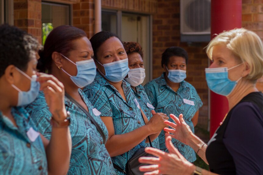 Aged care workers in uniform and wearing masks speak to a woman.