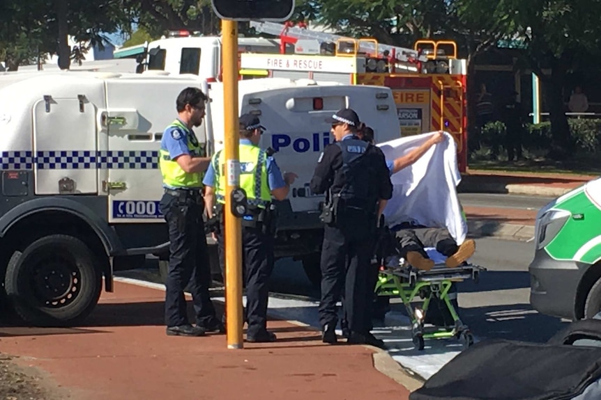 Three police officers stand next to a man on an ambulance stretcher near police vehicles with a fire truck in the background.