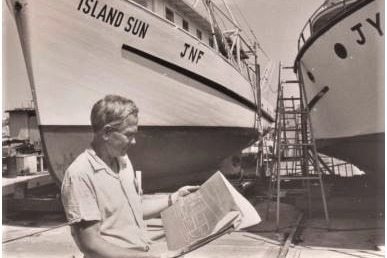Black and white photo of man looking at plans in front of two large white boats on land