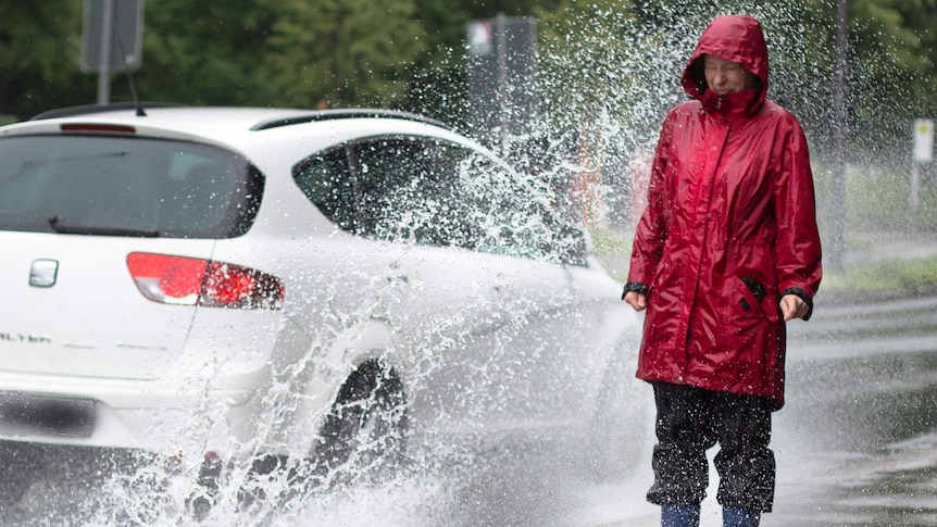 A woman, dressed in wet weather clothing, is splashed by the wheels of a passing car.