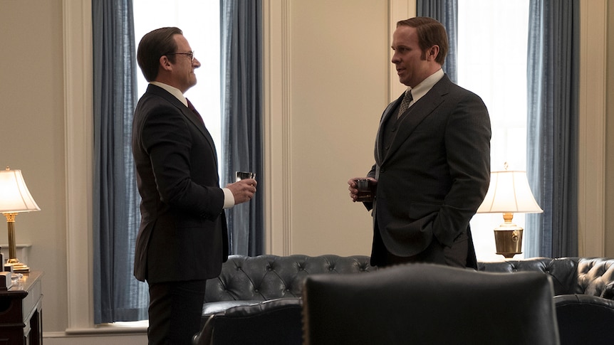 Colour still of Steve Carell and Christian Bale standing and talking with drinks in office in 2018 film Vice.