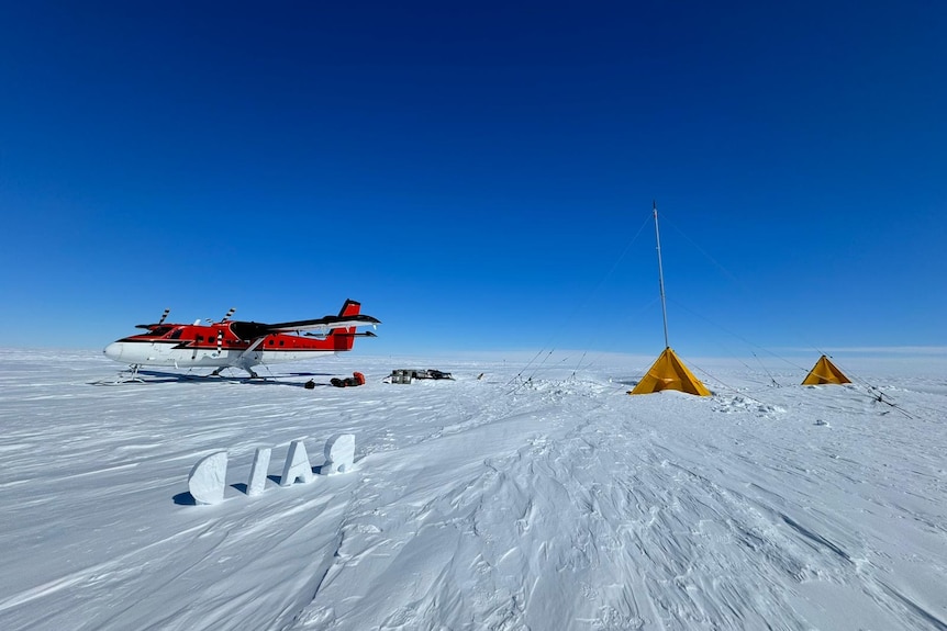 A red and white propeller plane sits on a white, snowy expanse. There are two yellow tents beside it.