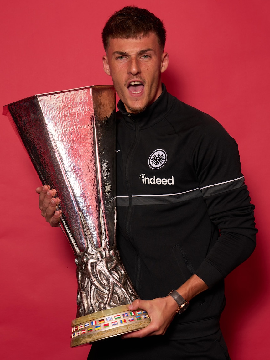 A male soccer player wearing a black jacket holds a big silver trophy in front of a red background