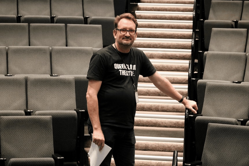 A man with scruffy hair poses in between rows of chairs in a theatre.