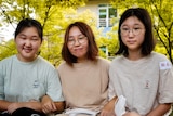 A young woman and her two pre-teen daughters sit on a garden bench. Trees with green-yellow leaves are in full bloom behind them