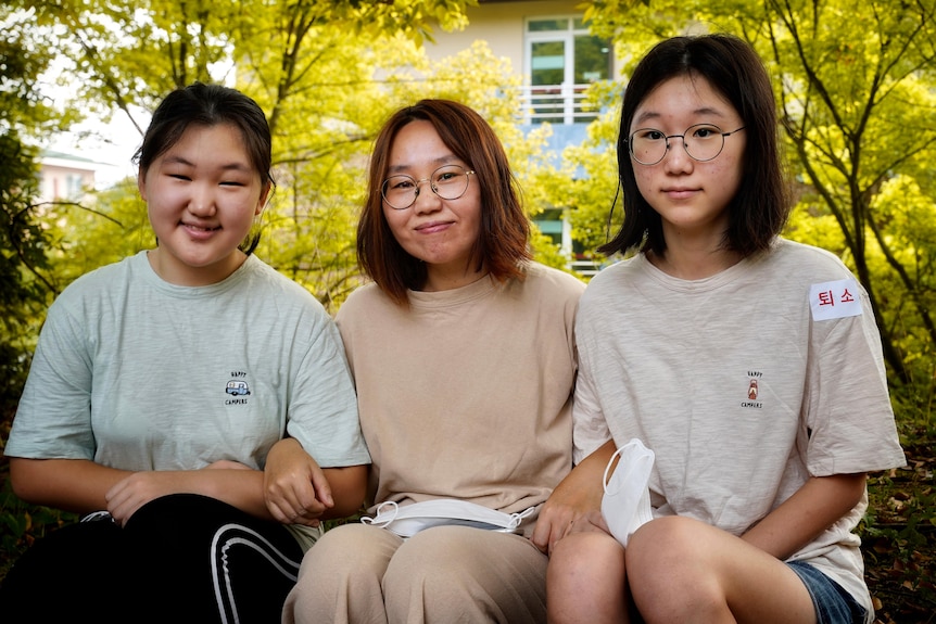 A young woman and her two pre-teen daughters sit on a garden bench. Trees with green-yellow leaves are in full bloom behind them