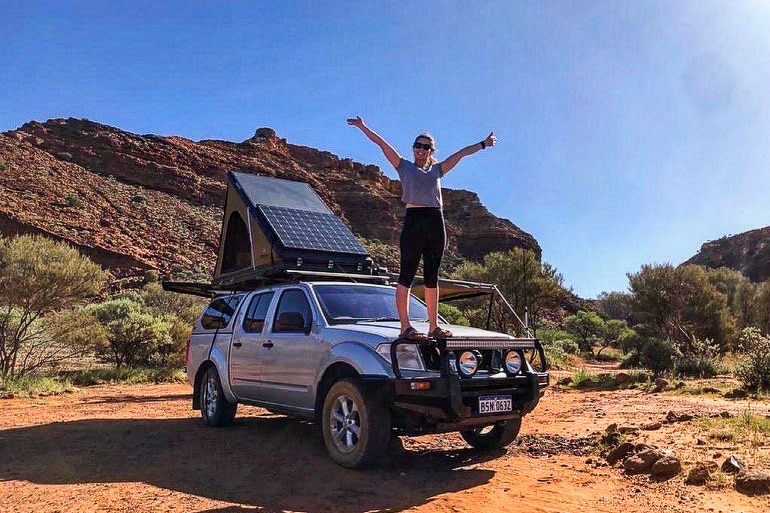 a woman stands on the roof of a four-wheel drive car with a solar panel on its roof