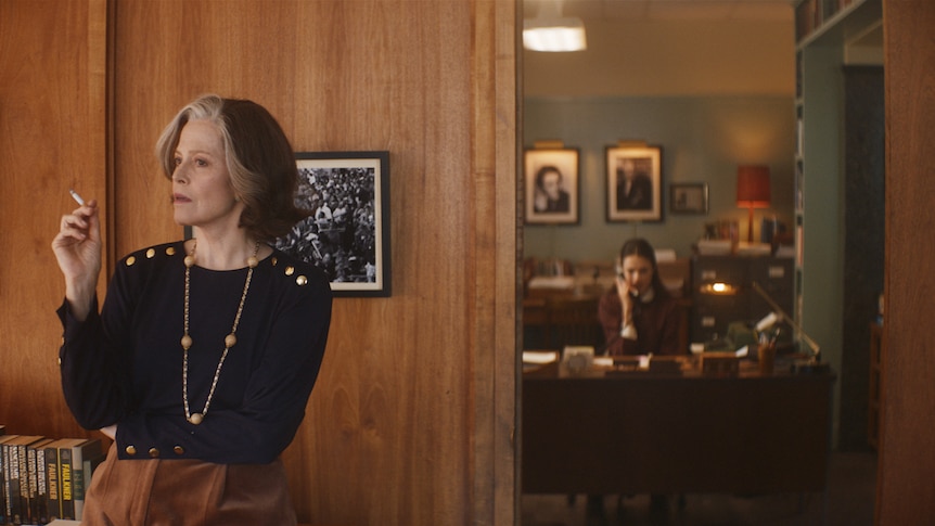 Interior office with Sigourney Weaver dressed stylishly, smoking, and Margaret Qualley in background, typing at desk.