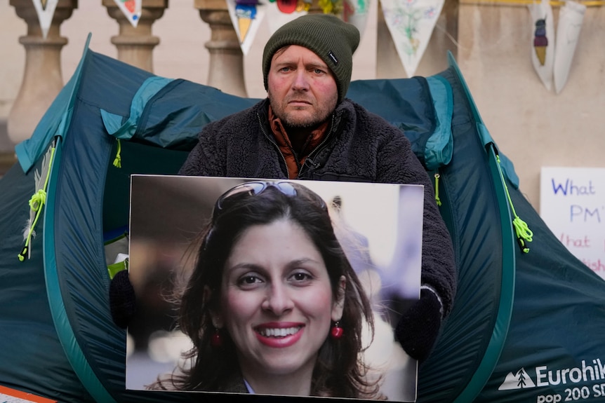 A man holds a picture of a smiling woman while sitting in front of tent.