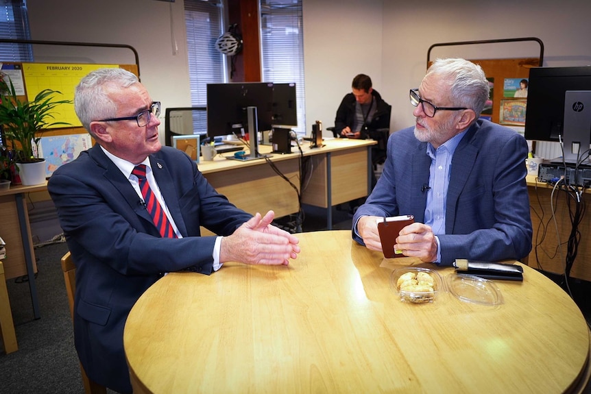 Two grey-haired men both wearing suits and glasses chat to each other in an office.