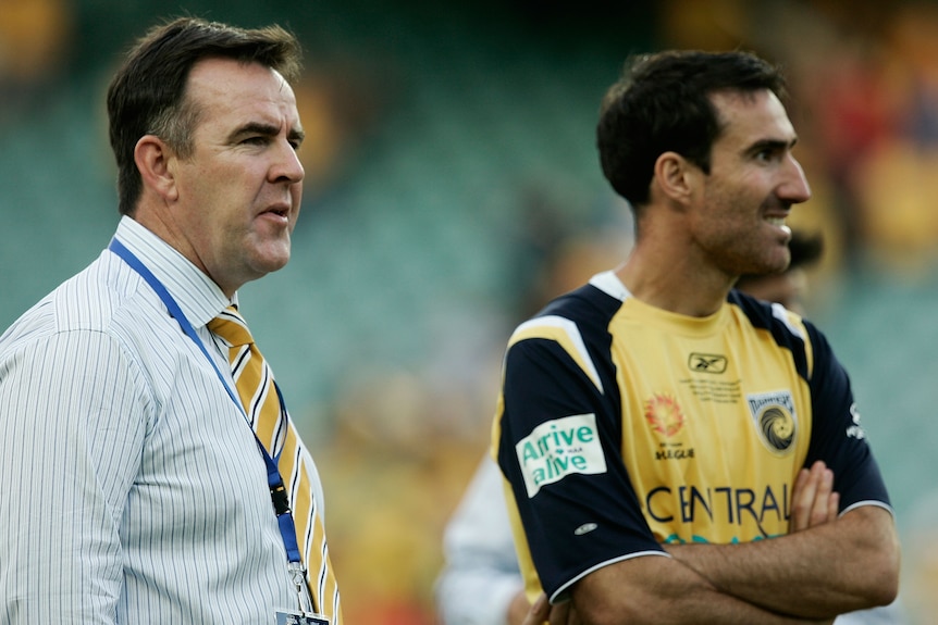 Two men, one wearing a collared shirt with a yellow tie and another wearing a yellow and blue shirt, stand next to each other