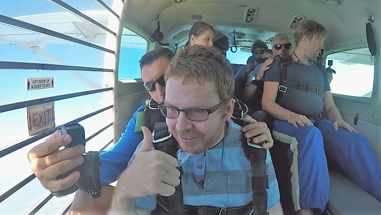 Man gives the thumbs up as he sits on a plane with other skydivers.