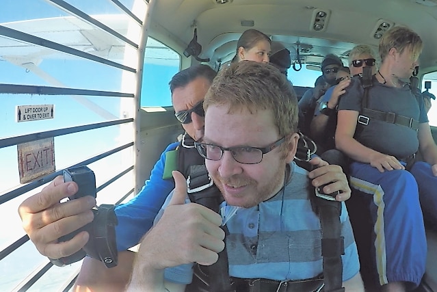 Man gives the thumbs up as he sits on a plane with other skydivers.