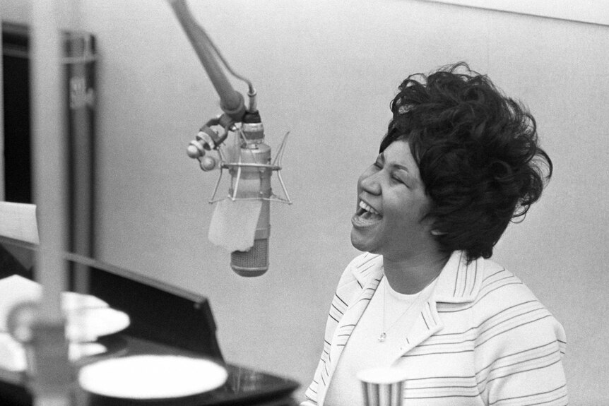 A black and white photo of Aretha Franklin singing into a microphone. She's wearing a white jacket with thin stripes.