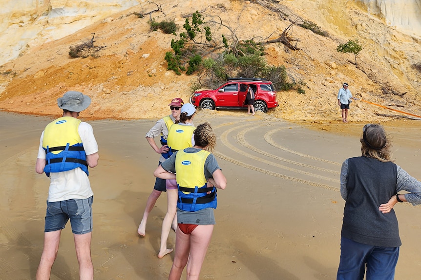Four people three in life vests look on as a red car is being dug out of the sand slip by two people