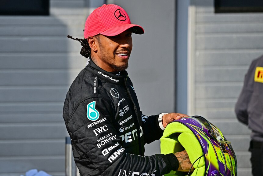 An F1 driver, holding his helmet and wearing a red cap, smiles after qualifying fastest.