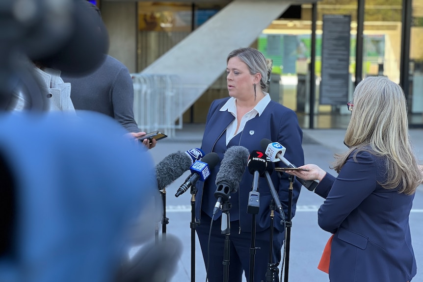 A woman with blonde hair stands in front of microphones and journalists outside a stadium.