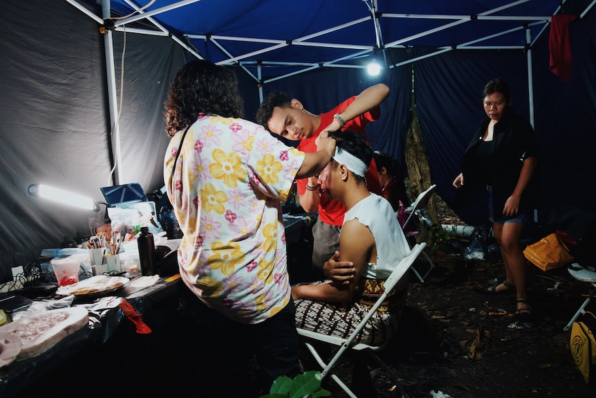 Makeup artists applying bloody makeup to an actor in a tent.