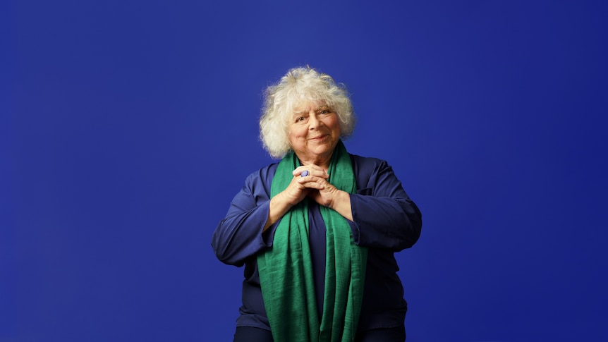 Miriam Margolyes, standing in front of a blue backdrop, with her hands clasped together, smiles at the camera.