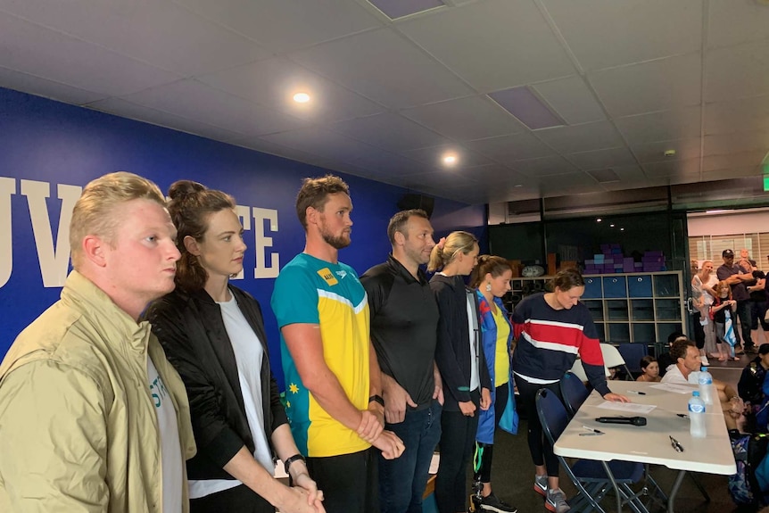 A group of athletes stand side by side in a room beside a table.