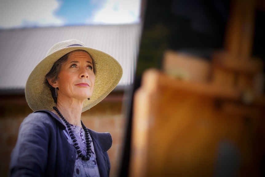 A woman in a wide brimmed hat looks up at an easel