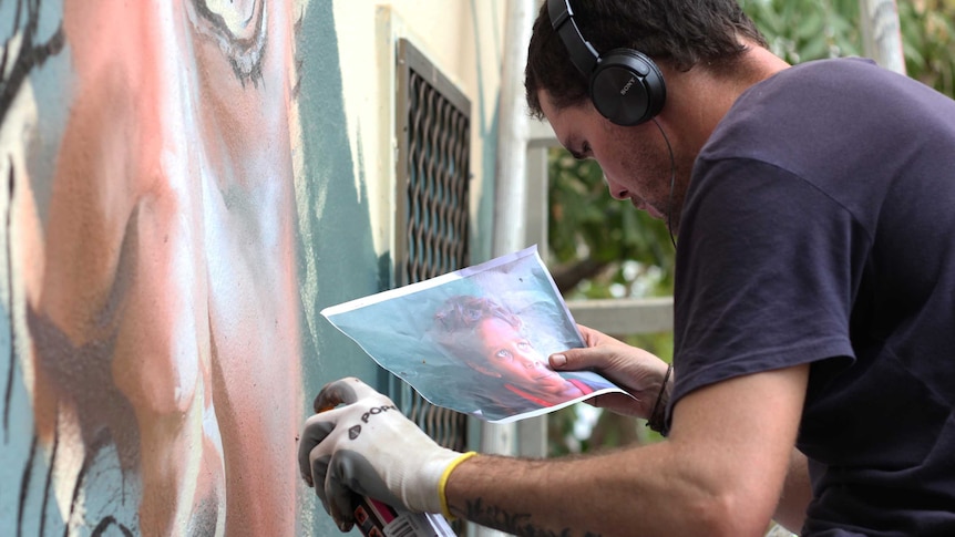 A man sitting on a plank up on a painted wall.