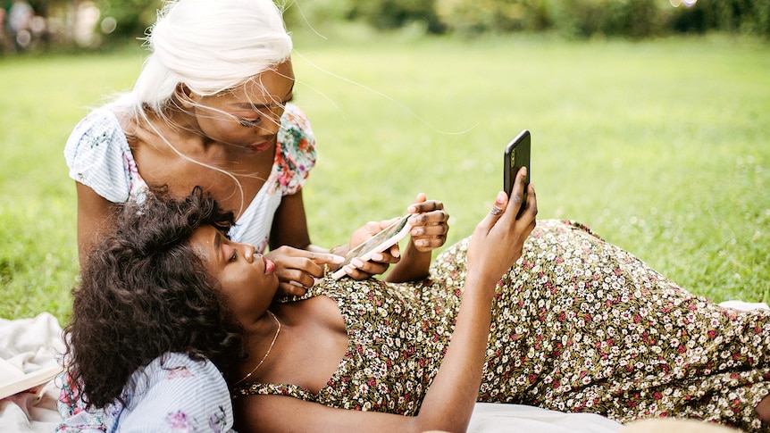 Two women are in a park on the grass on a rug, one sitting the other lying on her lap, and both are looking at their phones. 