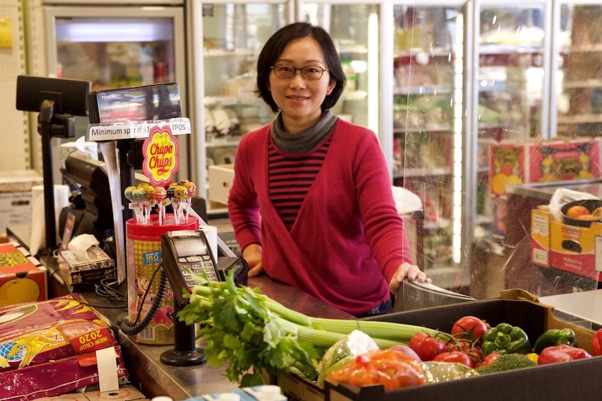 Wendy Yu stands behind the register at her grocery store, with a box of fruit and vegetables in front of her and fridges behind.
