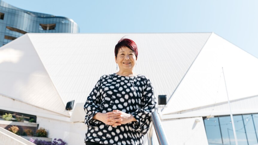 A 60-something Asian Australian woman stands in front of the Adelaide Festival Centre, her hands clasped in front of her