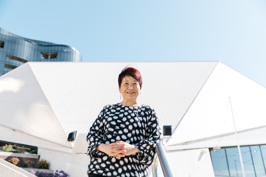 A 60-something Asian Australian woman stands in front of the Adelaide Festival Centre, her hands clasped in front of her
