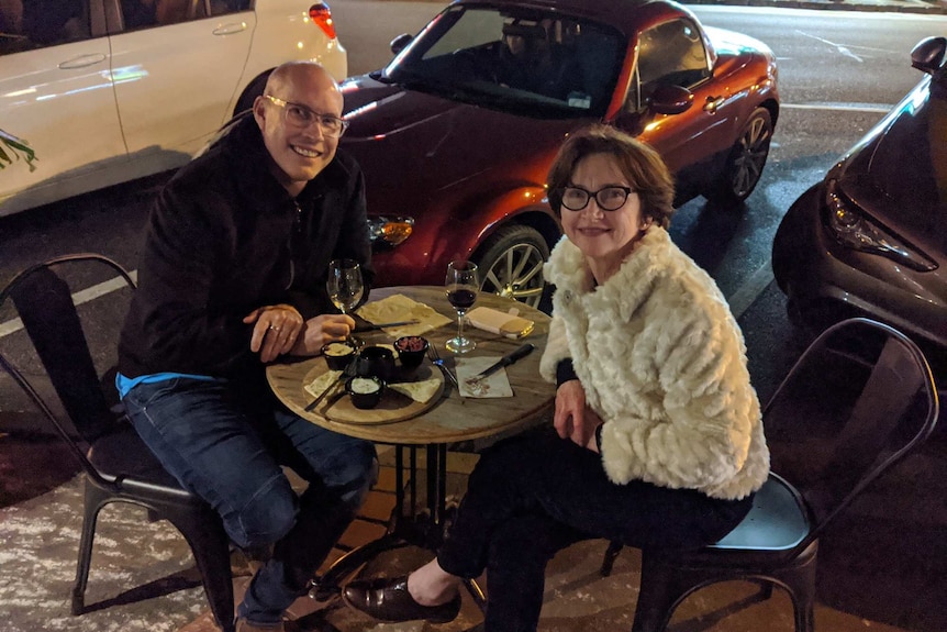 A man and woman sit at a table outside enjoying wine and nibbles.