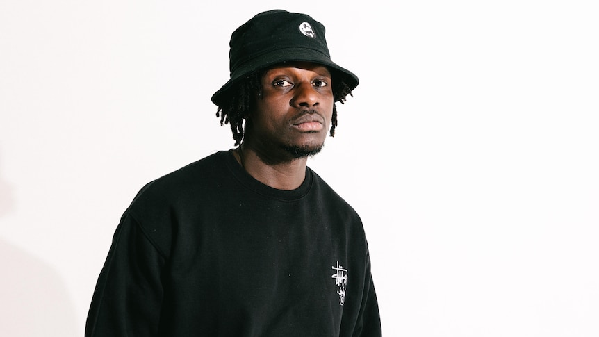 A man with dreadlocks wearing a black bucket hat looks neutrally at the camera