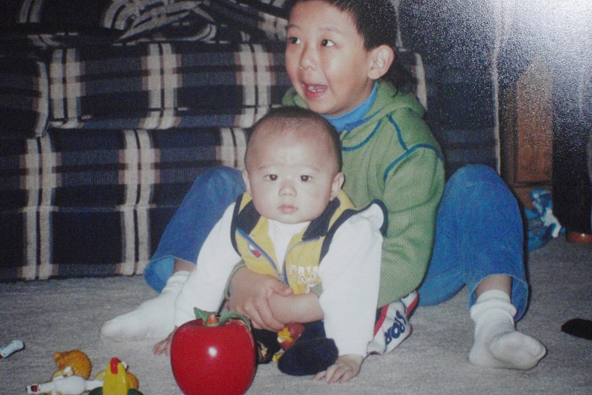 A young child holds a baby by the chest as they both crouch on the floor of what appears to be a lounge, a couch behind them.