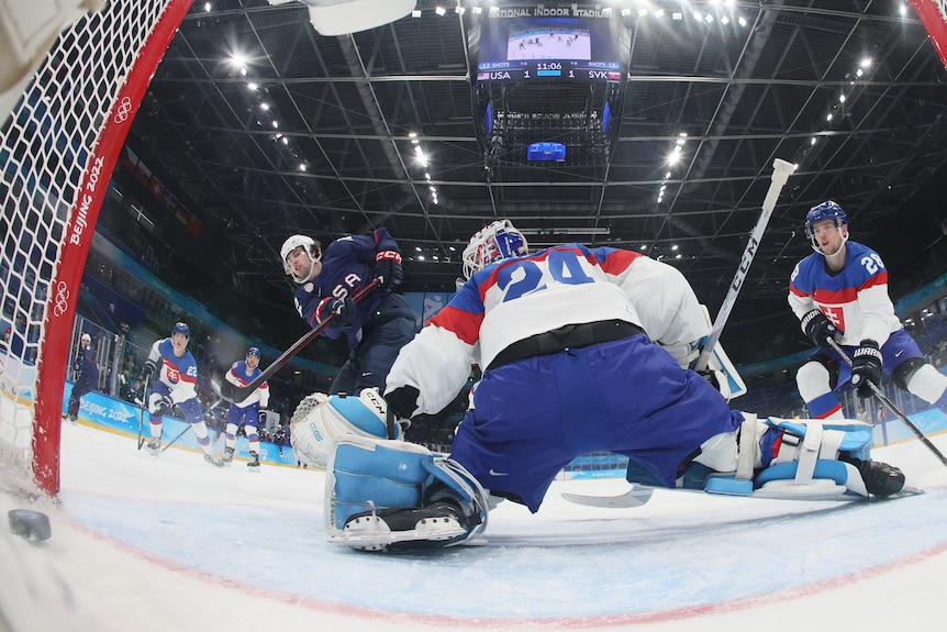 A shot from inside the ice hockey net shows the goaltender spreads himself as an attacker scores a goal.