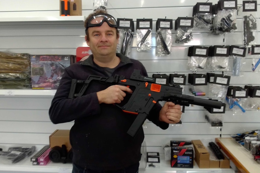 A man smiles holding a lifelike toy gun with safety goggles on his forehead.